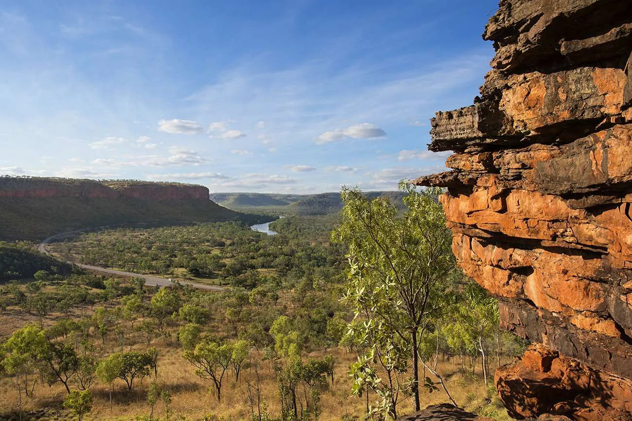 A scenic landscape photograph from Judbarra/Gregory National Park, showing a vast valley with green trees, a winding river, and distant cliffs under a blue sky, with a prominent orange-brown rock escarpment in the right foreground.