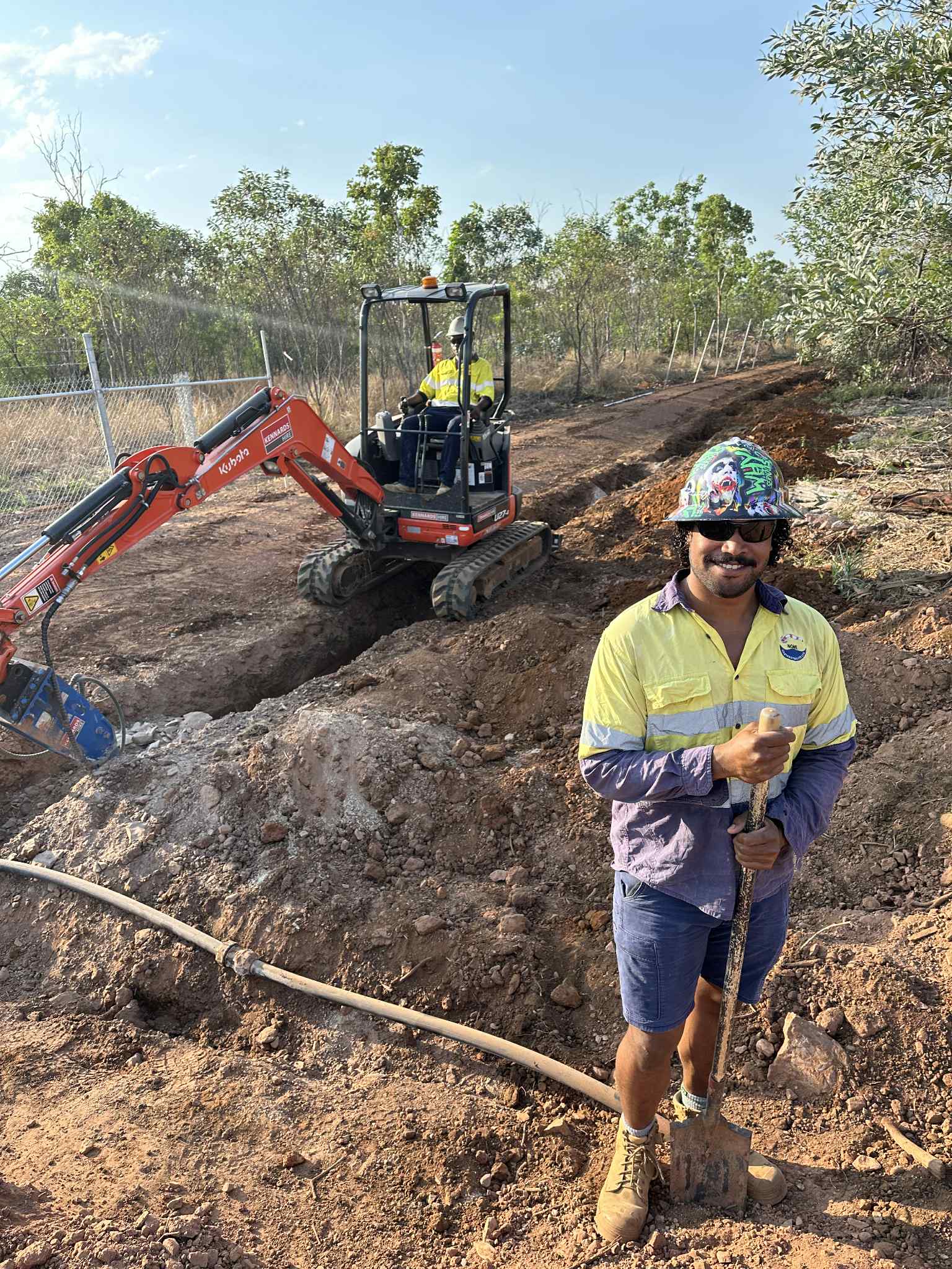 Person in high-visibility clothing and hard hat standing in a construction site with an excavator in the background.