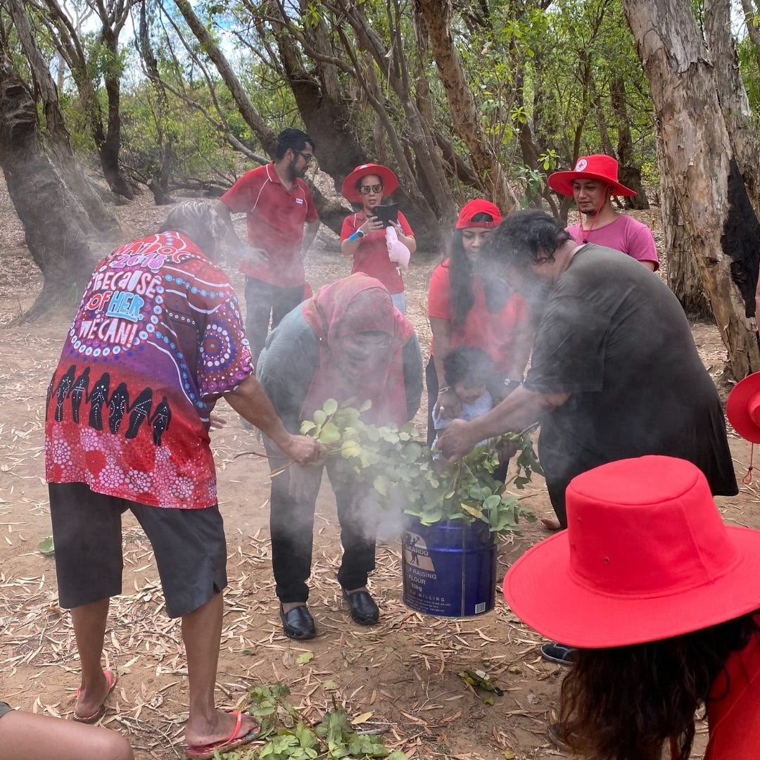 Group of people participating in a traditional smoking ceremony, performed by May Rosas, outdoors with trees in the background.